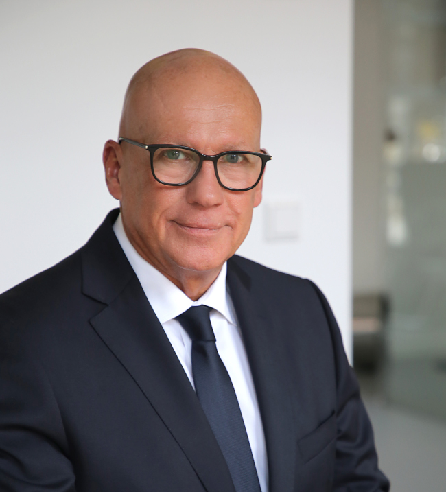 Ein gut gekleideter Mann mit Glatze und Brille, der in einem modernen, hellen Büro posiert.