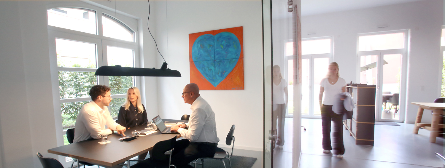 Ein modernes Büro mit drei Personen, die an einem Tisch sitzen, und einer weiteren Person, die im Hintergrund vorbeigeht. An der Wand hängt ein großes, buntes Herzgemälde.
