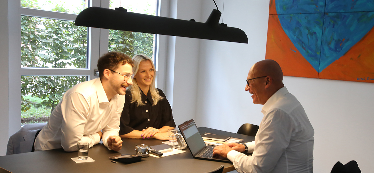 Drei Personen sitzen an einem Tisch in einem hellen Raum und unterhalten sich. Eine Frau und ein Mann schauen lächelnd zu einem weiteren Mann, der vor einem Laptop sitzt. An der Wand hängt ein großes, farbenfrohes Kunstwerk.
