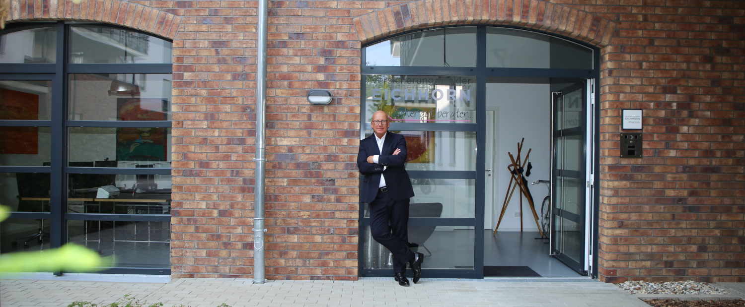 Ein Mann in einem Anzug steht vor einem modernen Bürogebäude mit einer Ziegelsteinfassade. Die Eingangstür ist offen, im Hintergrund sind Büromöbel zu sehen.