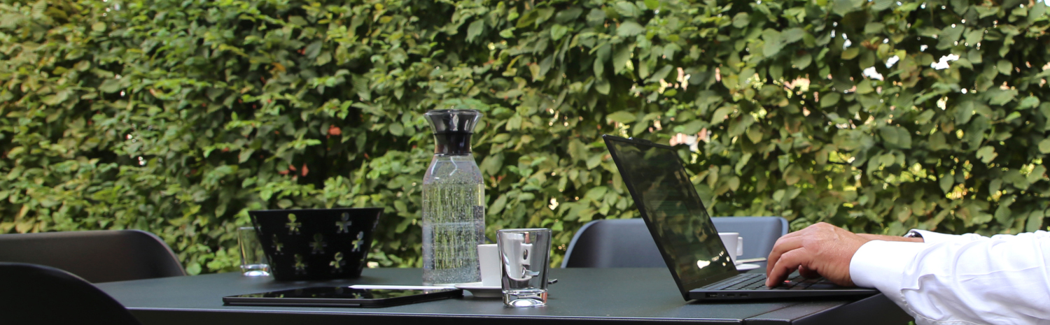 Ein Schreibtisch im Freien mit einem Laptop, einem Tablet, einem Glas Wasser und einer Kanne, umgeben von Pflanzen.
